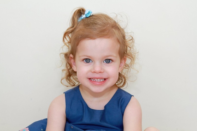 A little girl with pale skin, strawberry blonde hair and a blue dress smiles at the camera.